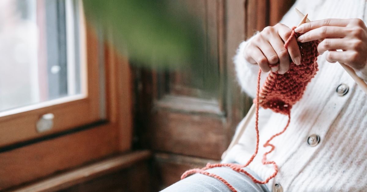 Crop faceless woman in warm wool cardigan sitting on chair near window and knitting with needles in daytime at home